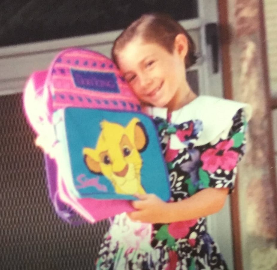 Picture of a young Sarah standing in a floral dress in front of her house holding a Lion King backpack and smiling at the camera.